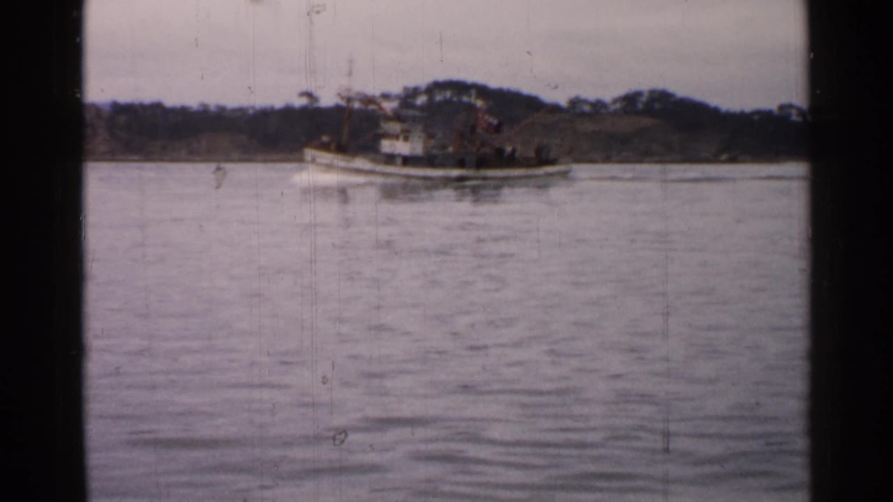 1952 Japan home-movie frame showing a white tugboat crossing calm water, with indistinct people on deck and a low wooded background.