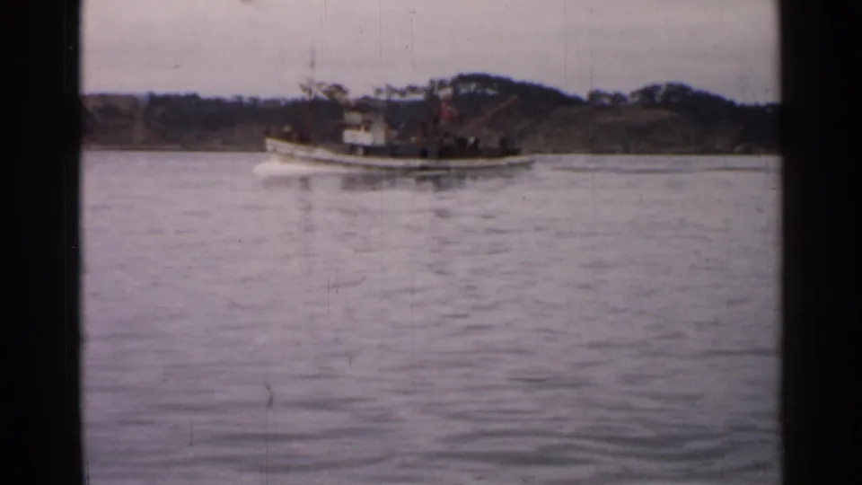 1952 Japan 8mm frame showing the tugboat nearing the right side of the shot, with calm water and a wooded shoreline behind it.