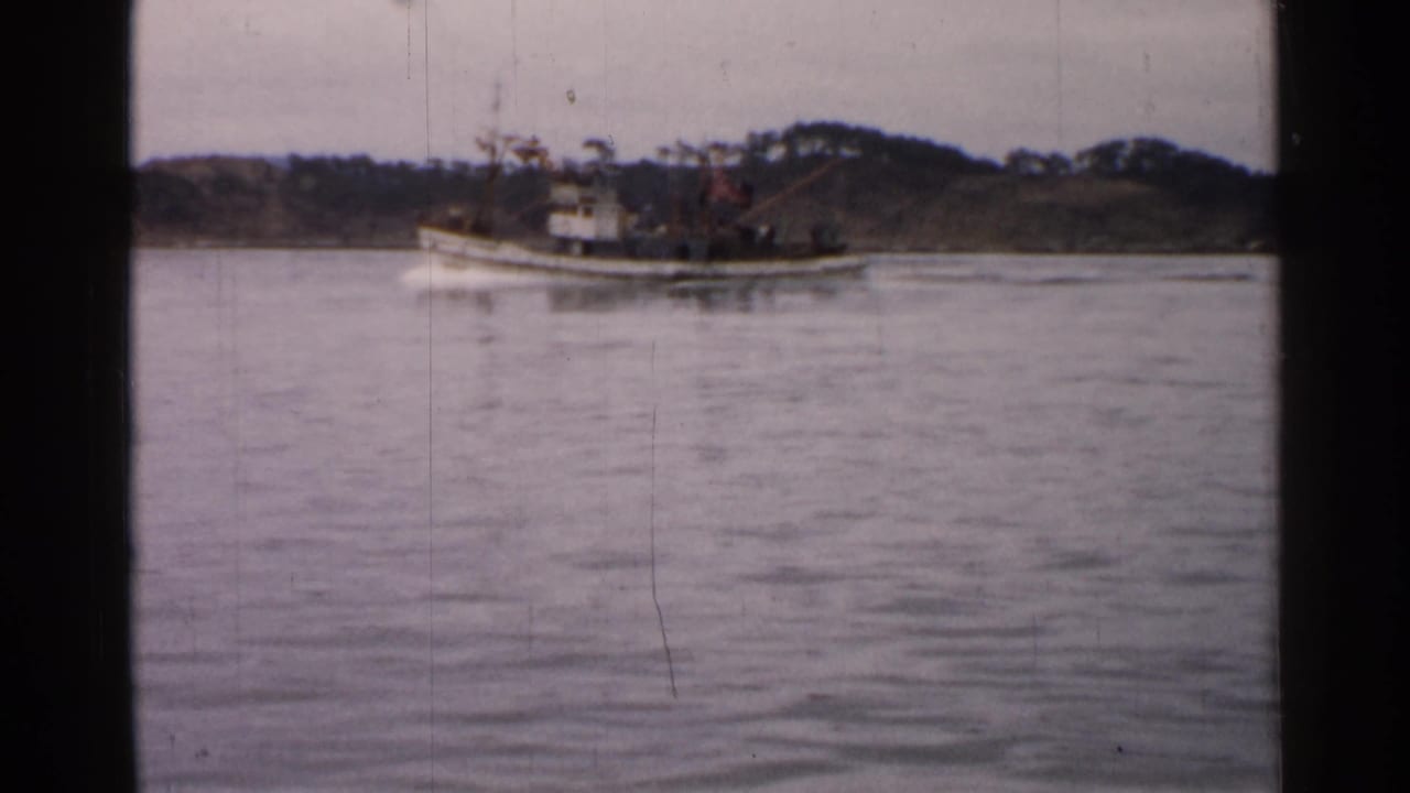 1952 Japan 8mm frame showing a small vessel moving across calm water near a low wooded shoreline.