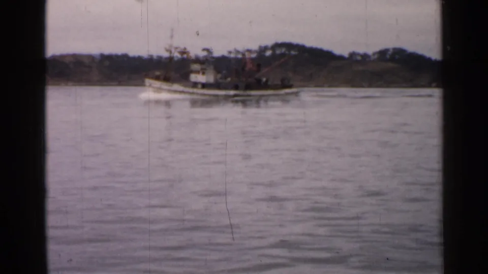 1952 Japan 8mm frame showing a small vessel moving across calm water near a low wooded shoreline.