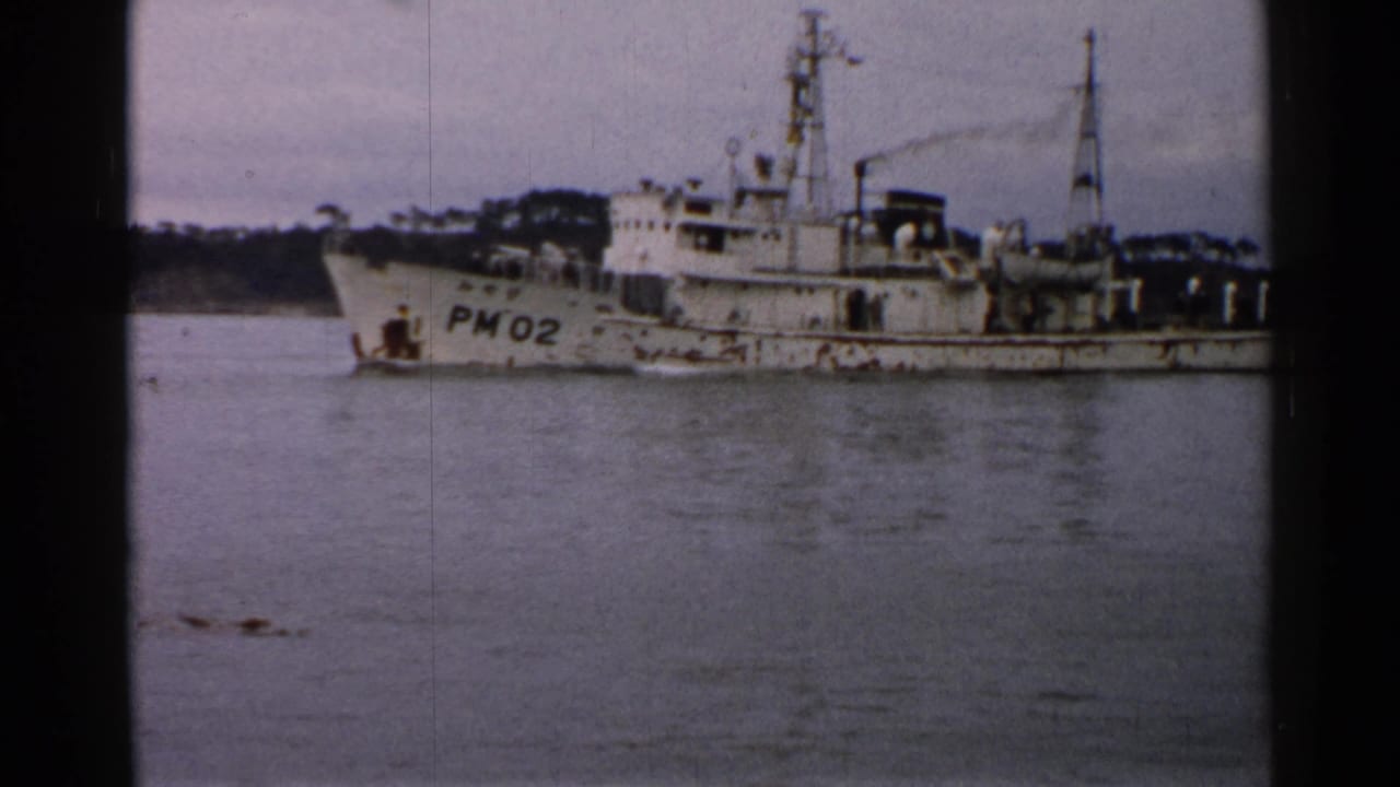 Aged 8mm frame from Japan showing the PM02 marked vessel on calm water with shoreline behind it.