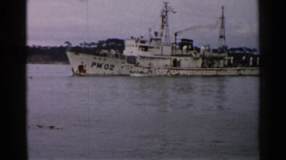 Aged 8mm frame from Japan showing the PM02 marked vessel on calm water with shoreline behind it.