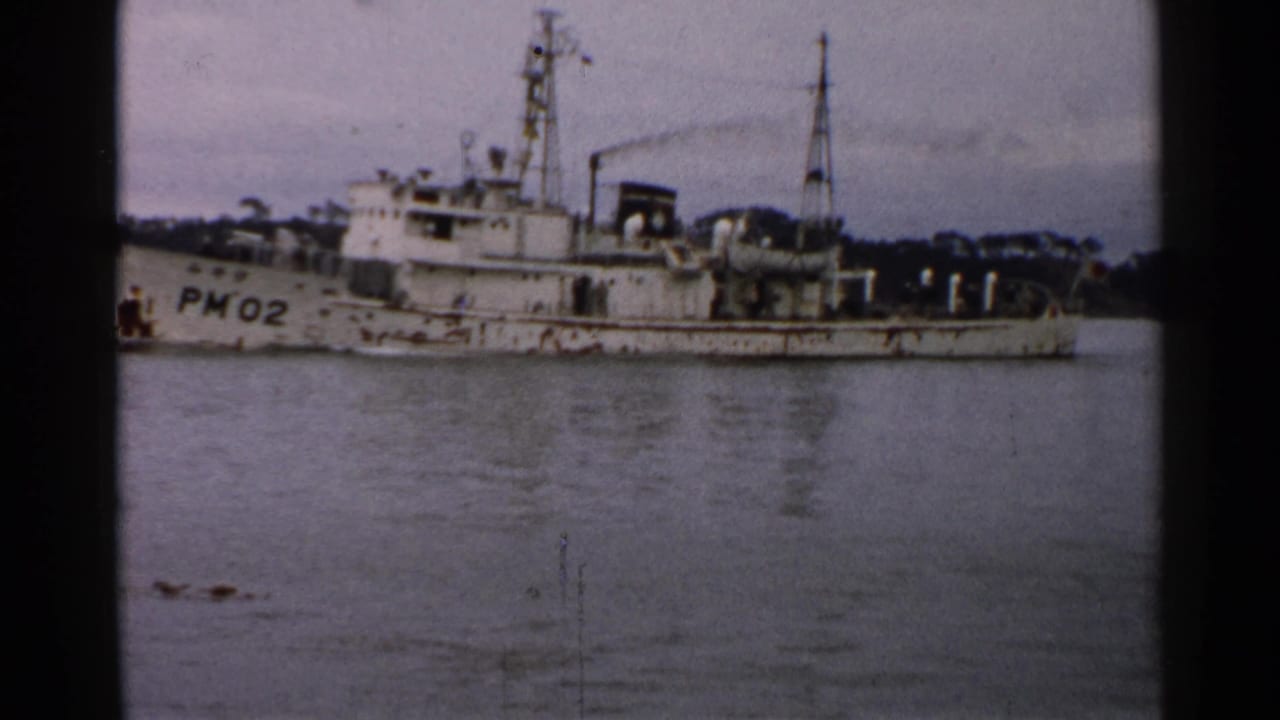 Broadside view of the PM02-marked vessel in 1952 Japan, with masts, deck gear, calm water, and distant trees visible.
