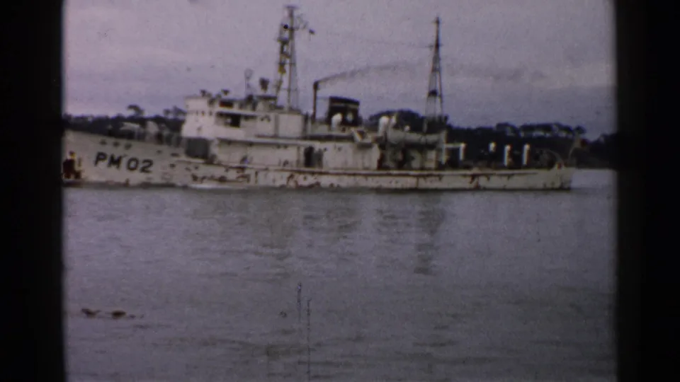 Broadside view of the PM02-marked vessel in 1952 Japan, with masts, deck gear, calm water, and distant trees visible.