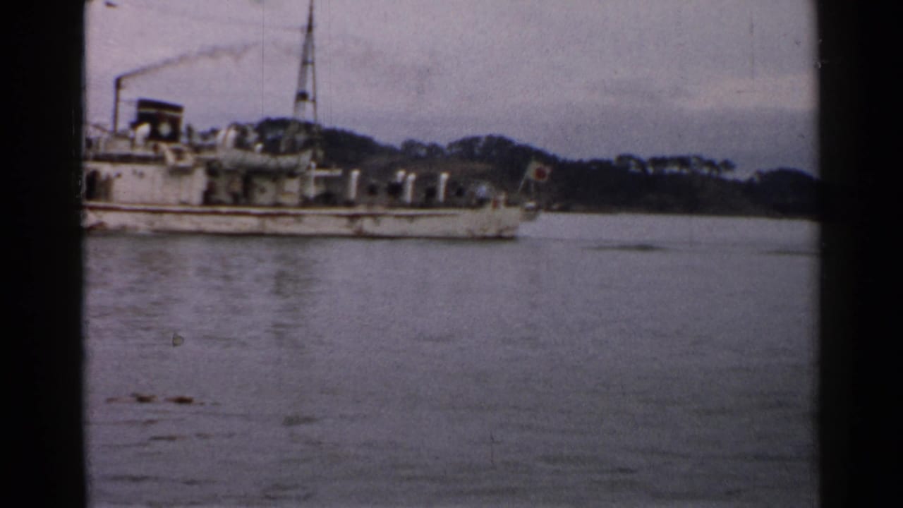 1952 Japan 8mm frame with a side view of a weathered vessel, Japanese stern flag, calm water, and low wooded shore.
