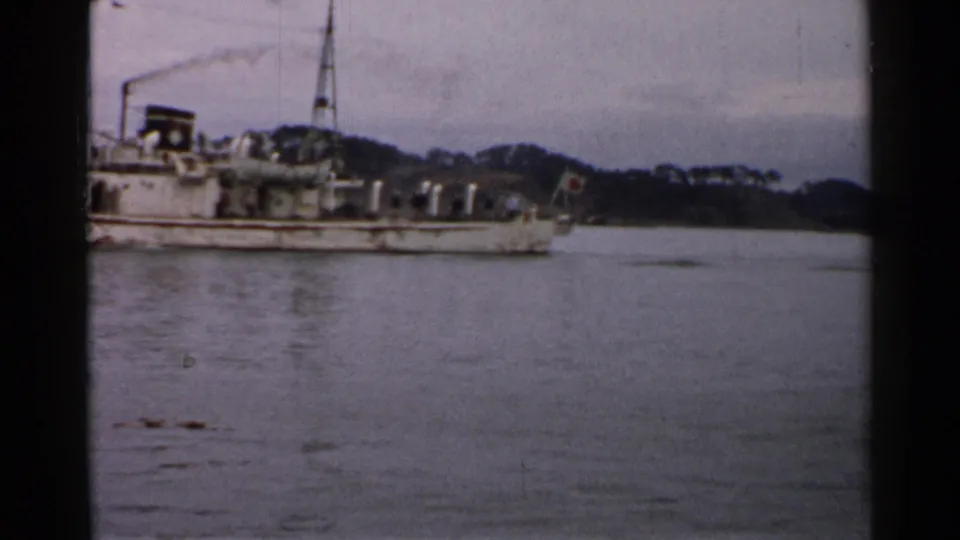 1952 Japan 8mm frame with a side view of a weathered vessel, Japanese stern flag, calm water, and low wooded shore.
