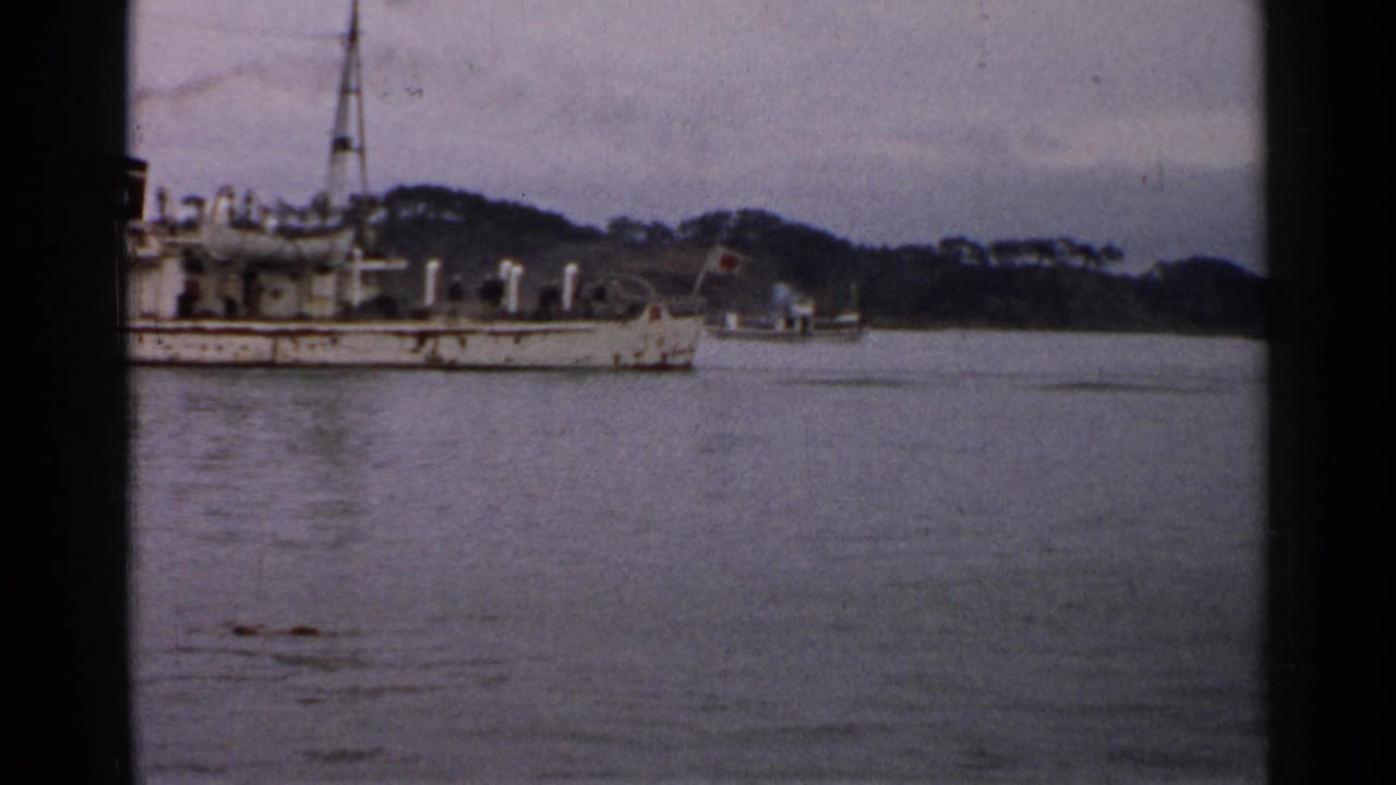 1952 Japan 8mm frame showing part of a white vessel along calm water, with a wooded shoreline and hills in the background.