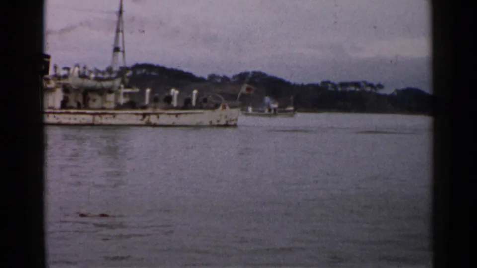 1952 Japan 8mm frame showing part of a white vessel along calm water, with a wooded shoreline and hills in the background.