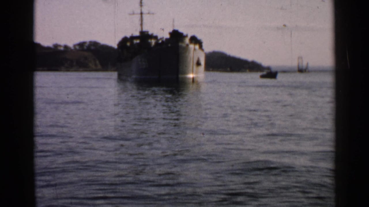 1952 Japan 8mm frame showing a darker bow-on view of a vessel on coastal water, with small distant boats and hills beyond.