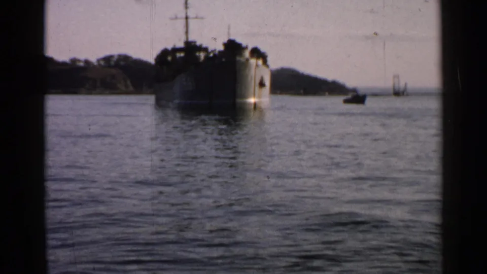 1952 Japan 8mm frame showing a darker bow-on view of a vessel on coastal water, with small distant boats and hills beyond.