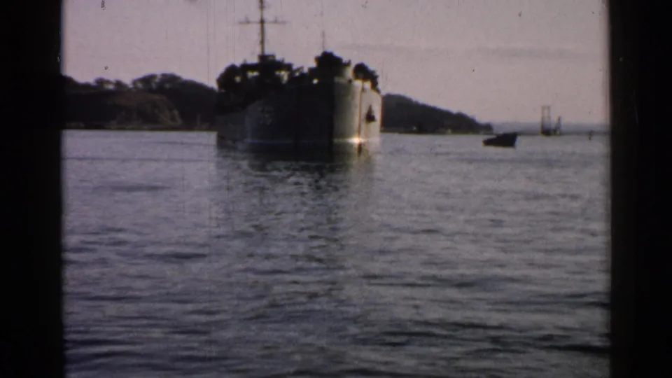 1952 Japan 8mm frame showing a large naval vessel offshore, calm coastal water, a wooded hill, and dark film-frame edges.