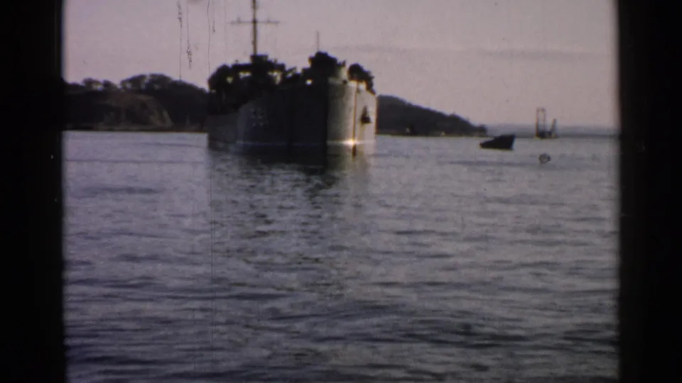 Distant 1952 Japan coastal view of a large ship at anchor, with small boats, tree-covered land, and visible 8mm frame borders.