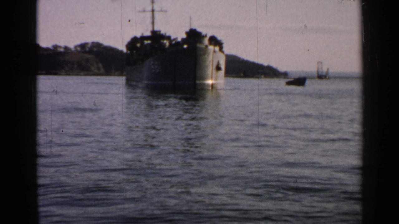 Soft 8mm frame of a large naval vessel off the coast in Japan, with calm water, a wooded shoreline, and heavy gate-edge framing.