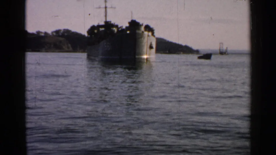 Soft 8mm frame of a large naval vessel off the coast in Japan, with calm water, a wooded shoreline, and heavy gate-edge framing.