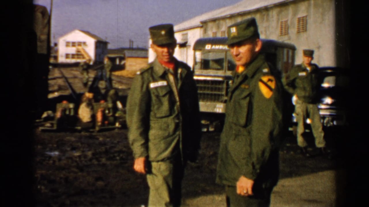 1952 Japan 8mm frame showing two soldiers in olive drab uniforms beside military vehicles and low buildings in a muddy compound.
