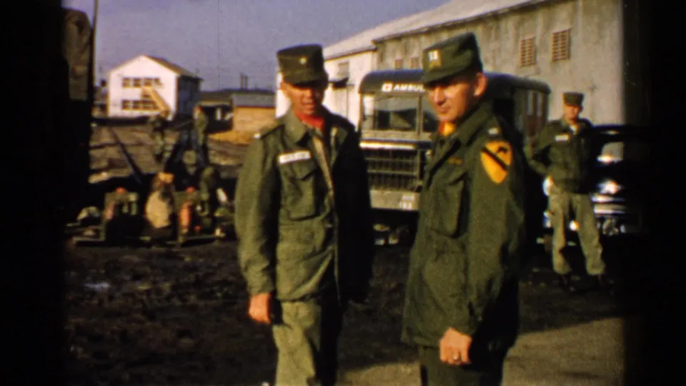 1952 Japan 8mm frame showing two soldiers in olive drab uniforms beside military vehicles and low buildings in a muddy compound.