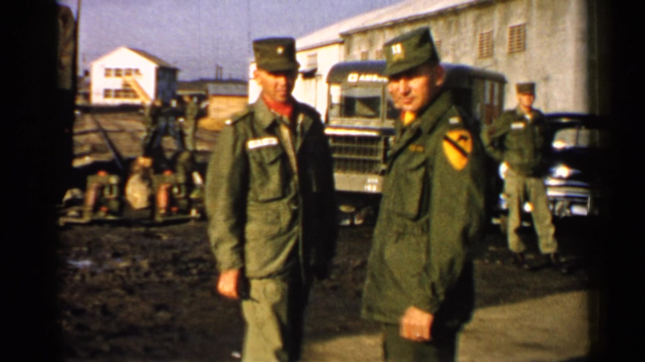 Two U.S. Army soldiers stand near a military truck and parked car in a muddy base yard.