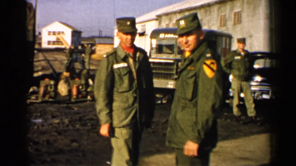 1952 Japan 8mm frame of uniformed U.S. Army soldiers talking beside a truck, car, base buildings, and equipment.