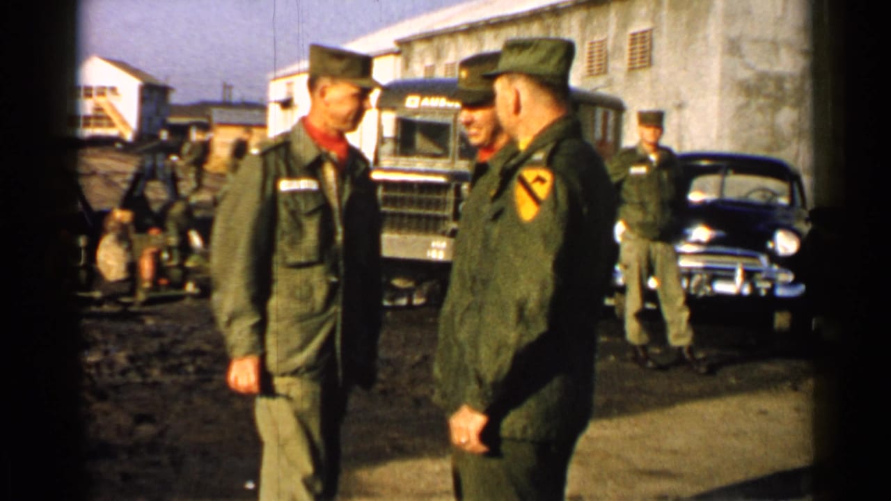 Three U.S. Army soldiers in 1952 Japan stand in conversation near a marked truck, parked car, and base structures.