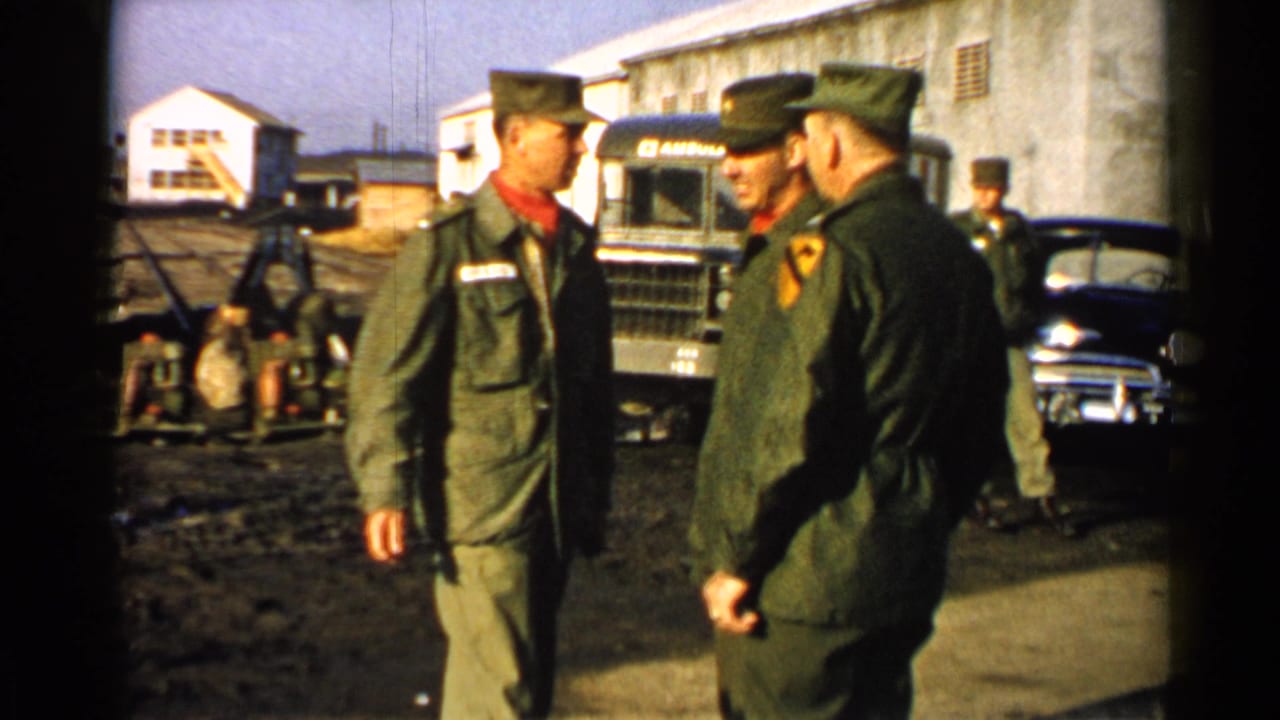 1952 Japan home-movie frame of Army soldiers conversing in a muddy base yard, with another serviceman passing behind them.