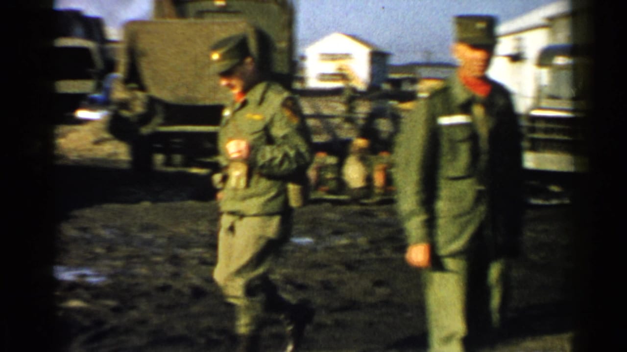 Two soldiers walk across muddy ground with canvas-covered military trucks behind them.