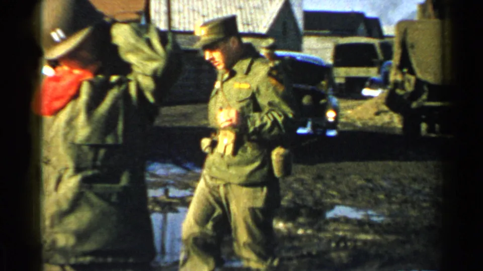 A soldier in olive uniform walks near another serviceman, parked vehicles, and puddles in the 1952 Japan military compound footage.