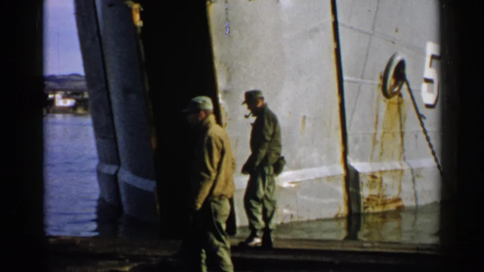Two uniformed men appear near a weathered white hull in Japan 1952 footage, with one partly in shadow and water behind them.