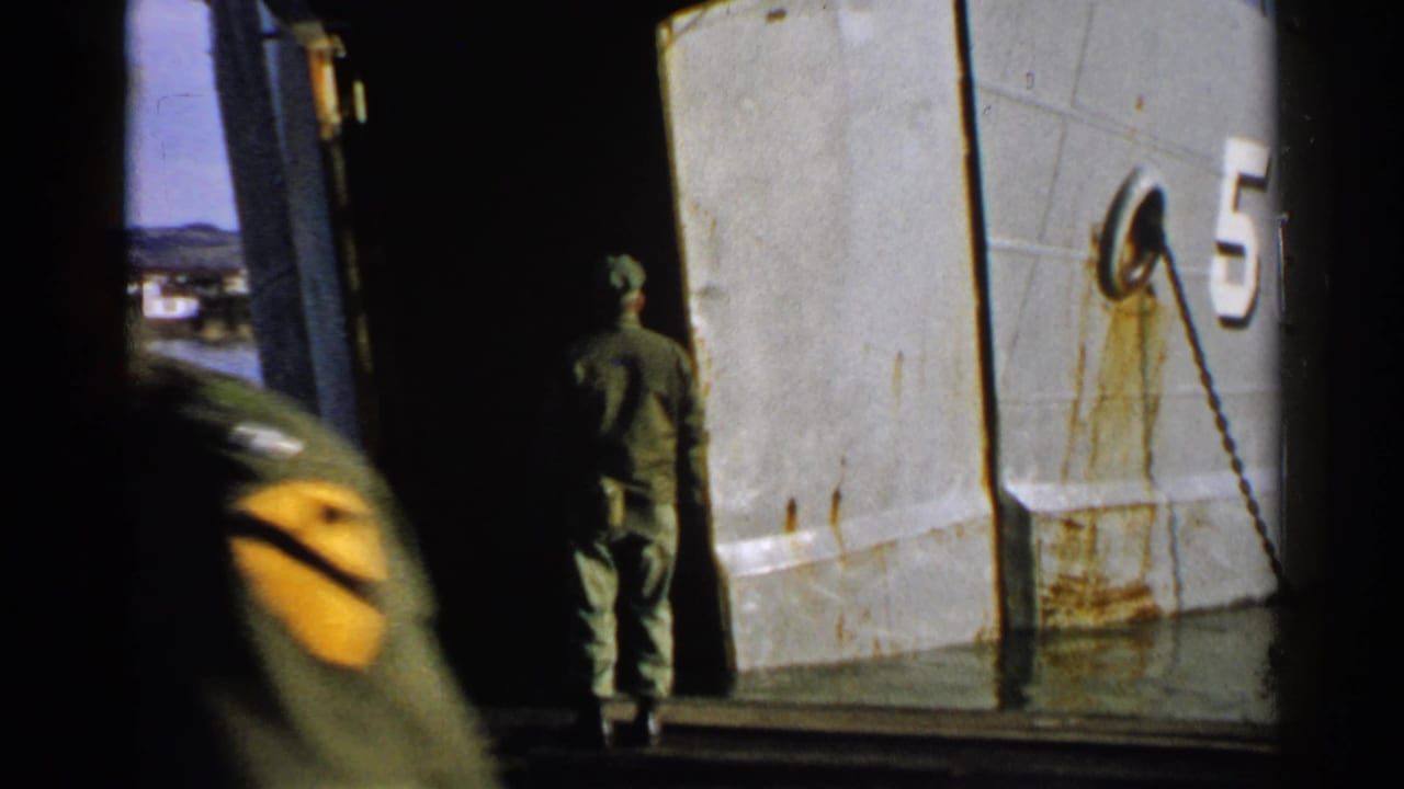 A uniformed man faces the open ramp of a gray vessel while another service member moves through the foreground.