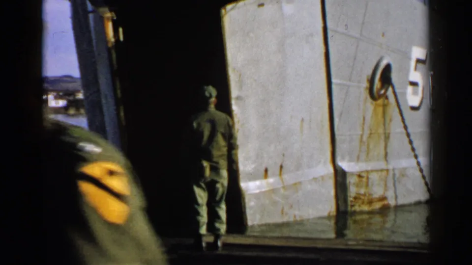 A uniformed man faces the open ramp of a gray vessel while another service member moves through the foreground.