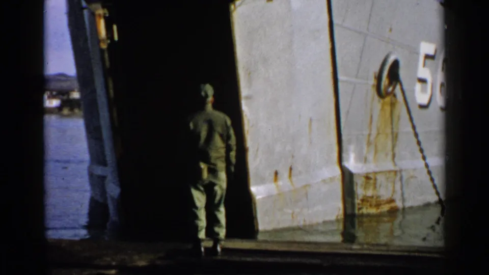 The man in green uniform walks toward the dark ramp opening of a gray vessel, with part of the hull number visible.