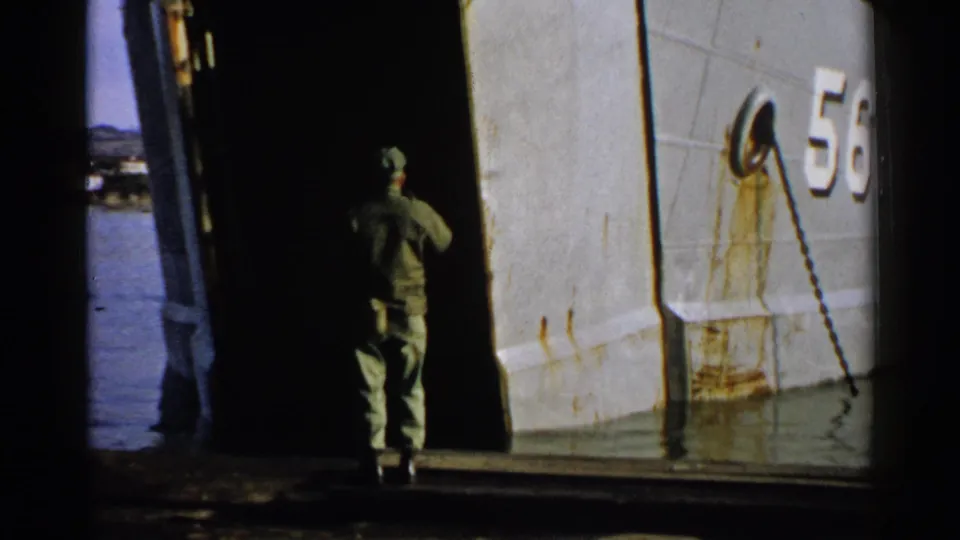 In the final frame, the uniformed man stands near the open ramp beside the gray hull marked 56 and a hanging chain or cable.