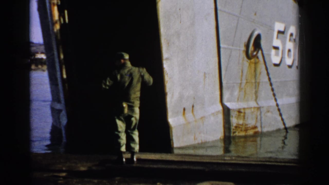 Uniformed man signals beside an open bow ramp on a ship hull numbered 561.