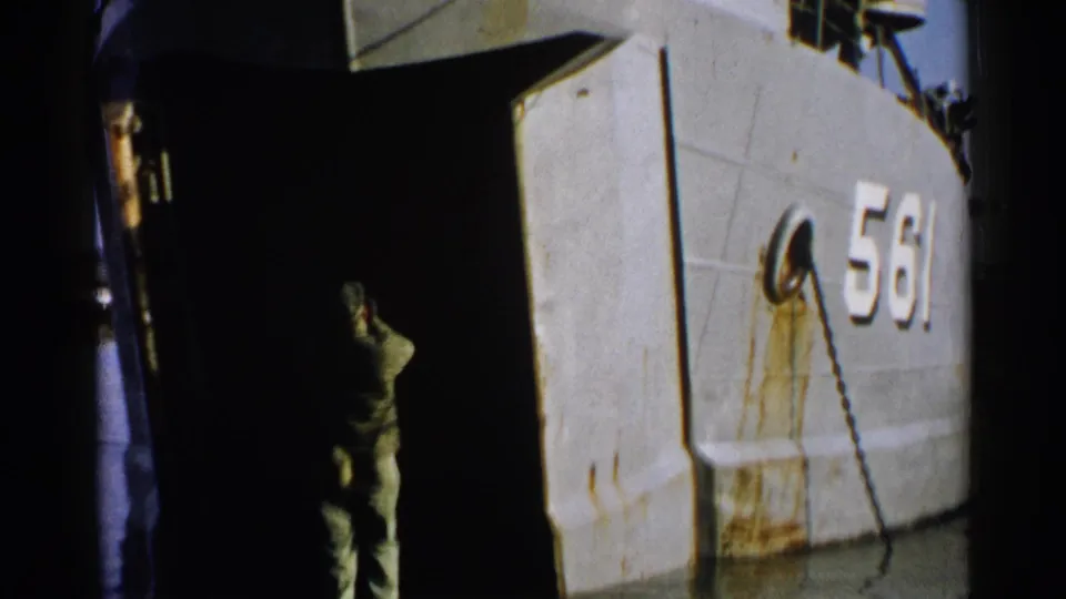 A uniformed man at the dock faces the open ramp of the numbered vessel, with a chain fitting and rust-stained hull panels visible.