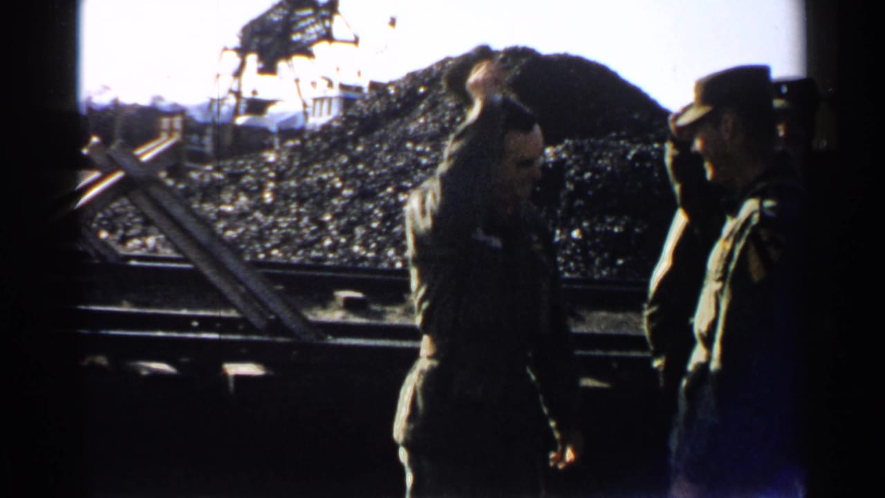 Two uniformed men stand near rail tracks and a large coal pile.