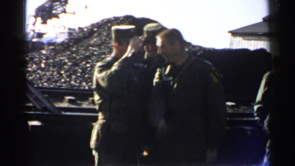 1952 Japan 8mm frame showing uniformed men gathered in a coal yard, one tipping his cap near rail tracks.