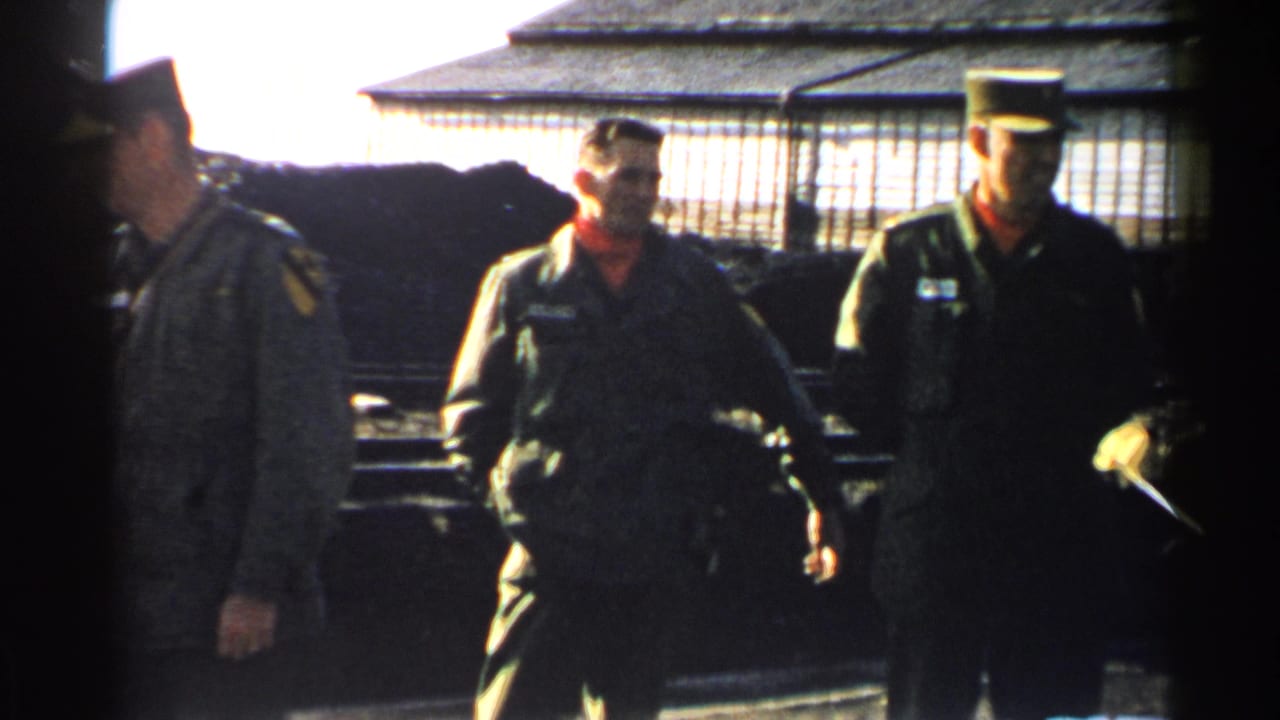 Two military men in green uniforms, one with a peaked cap and red scarf, move past a wire enclosure in Japan, 1952.