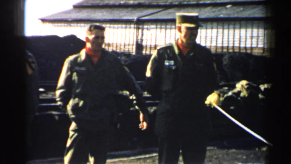 Two U.S. military men in green uniforms stand near a wire enclosure, with a long stick or baton entering the frame.