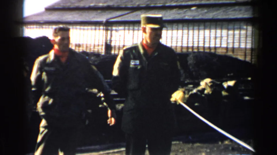 Two uniformed military men stand near a wire-fenced enclosure in Japan, with sunlit base structures behind them.