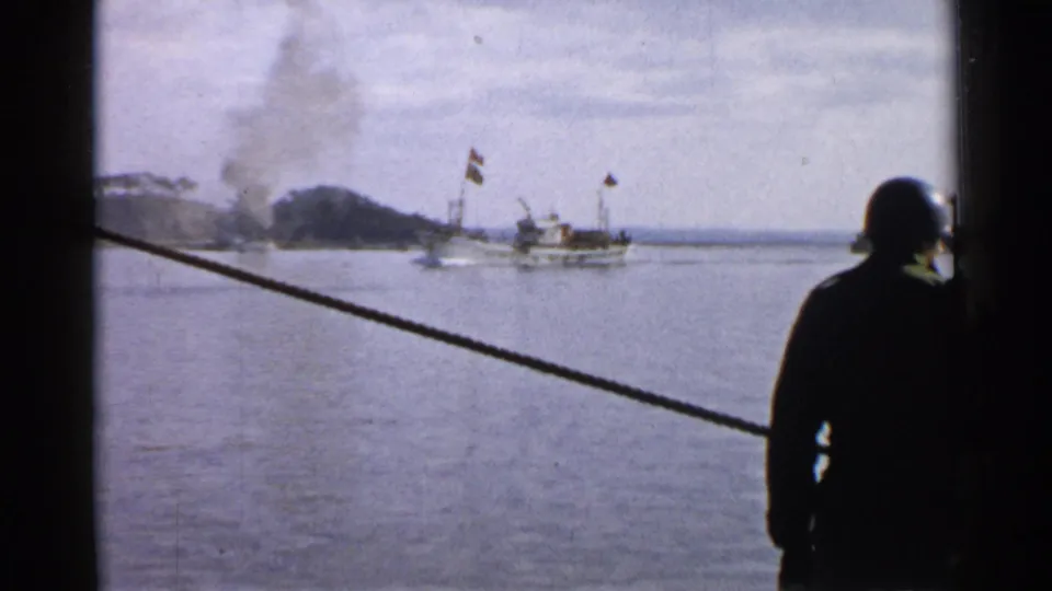 Viewed from a ship window in Japan 1952, a small harbor boat passes near shoreline smoke with a silhouetted figure at right.