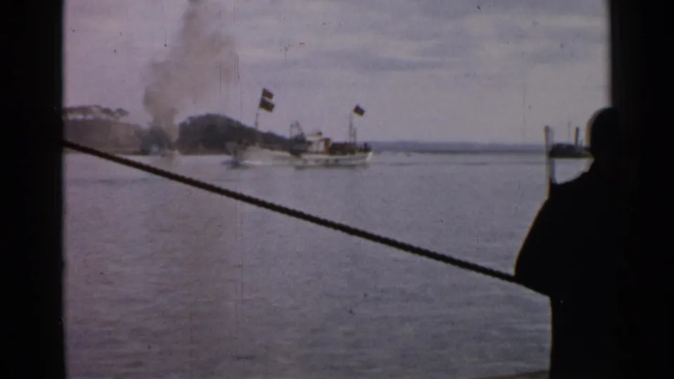 1952 Japan 8mm frame showing a smoking ship at sea, distant shoreline, mast flags, and a diagonal rope from the filming vessel.