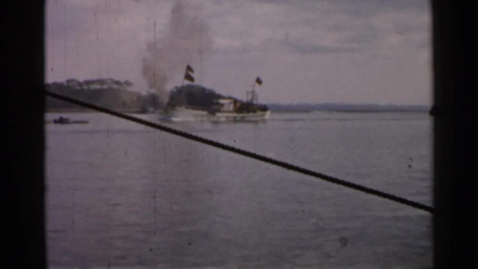 1952 Japan 8mm frame showing a smoking vessel, flags, calm sea, and a low distant shoreline viewed from another boat.