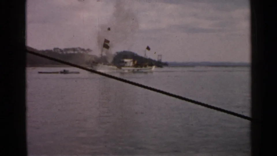 1952 Japan 8mm frame showing a distant steam cargo vessel crossing calm coastal water below a wooded shoreline.