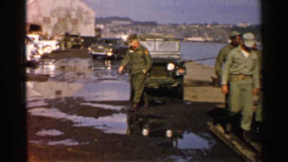 1952 Japan 8mm frame of soldiers on a wet waterfront dock with a military jeep, parked cars, a large building, water, and hills behind them.