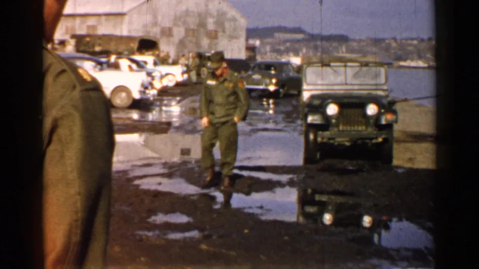 1952 Japan waterfront base scene with a soldier walking on a puddled dock near a jeep, parked cars, water, and a large building.