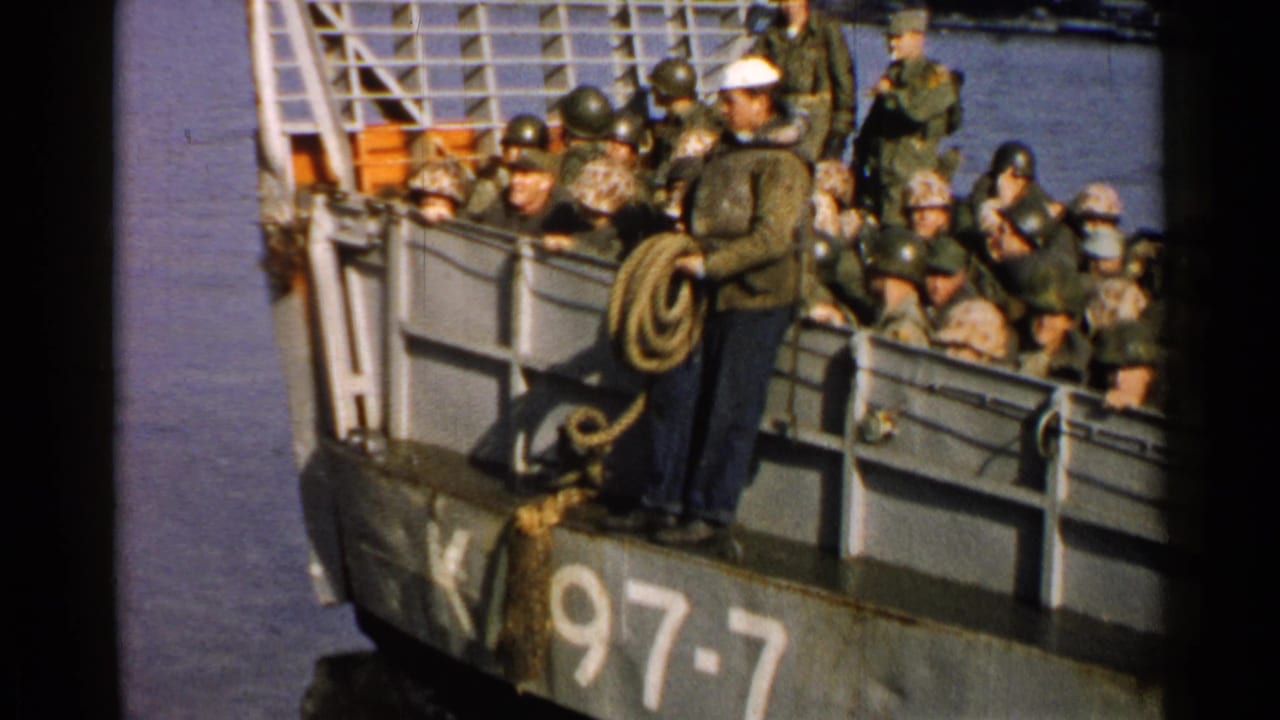 Helmeted soldiers crowded on a gray ferry as a sailor handles a coiled rope.