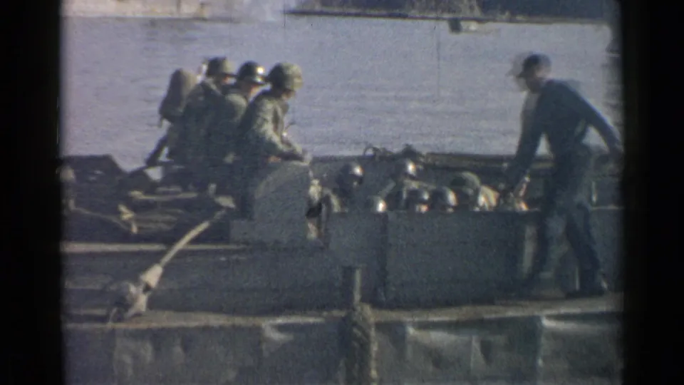 1952 Japan 8mm frame showing a standing crewman or soldier near seated helmeted troops on a ferry crossing water.