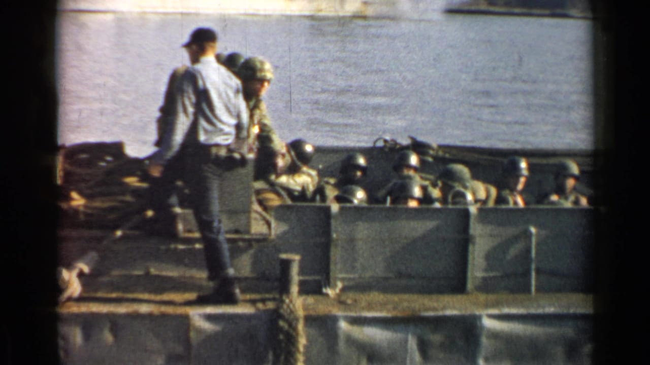 Helmeted soldiers sit in a landing craft beside calm water as a standing man faces them.
