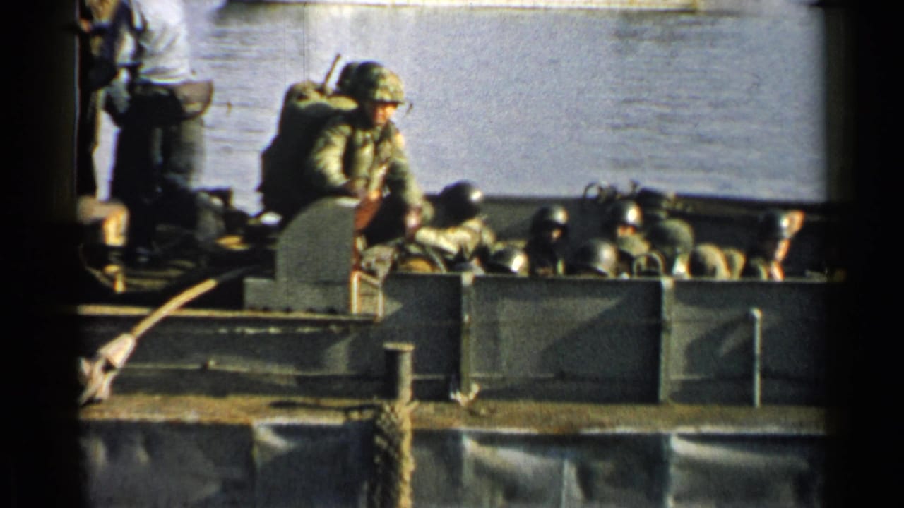 Soldiers in helmets sit low inside a landing craft with water behind them.