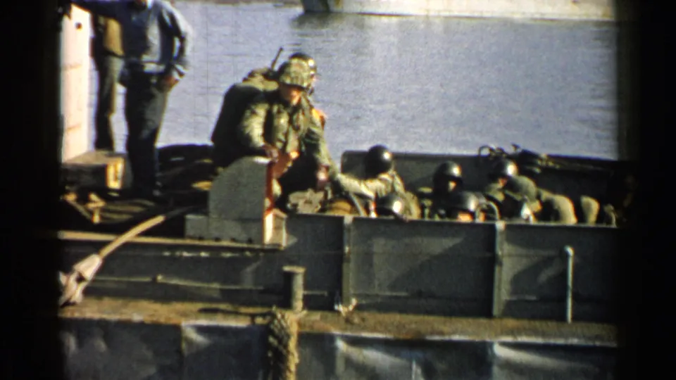 1952 Japan 8mm frame showing a soldier with a field pack near seated helmeted troops in a landing craft beside dock ropes and water.