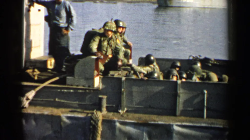 1952 Japan 8mm frame of U.S. soldiers in helmets and packs gathered in a landing craft, with a standing man, dock rope, and calm water behind.